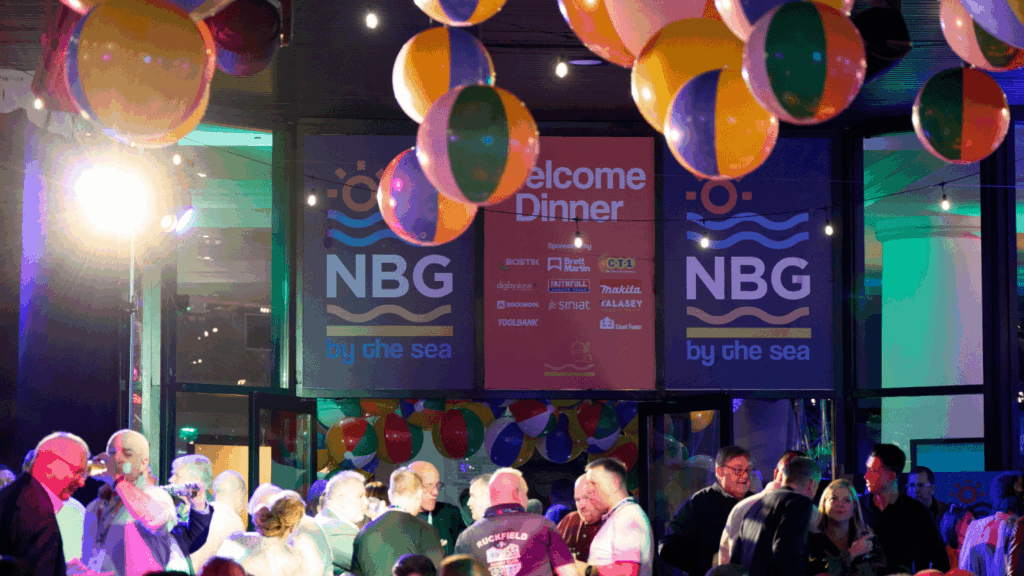 A lively indoor conference & exhibition event with people socializing under colorful beach balls and lights. A large screen displays “Welcome Dinner NBG by the sea.” The atmosphere is festive and vibrant.