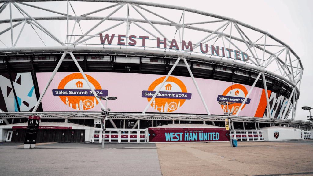 The exterior of West Ham United’s stadium displays large digital billboards advertising the London Conference: Sales Summit 2024 event. The stadium’s name is visible above the entrance, and the area outside looks empty.