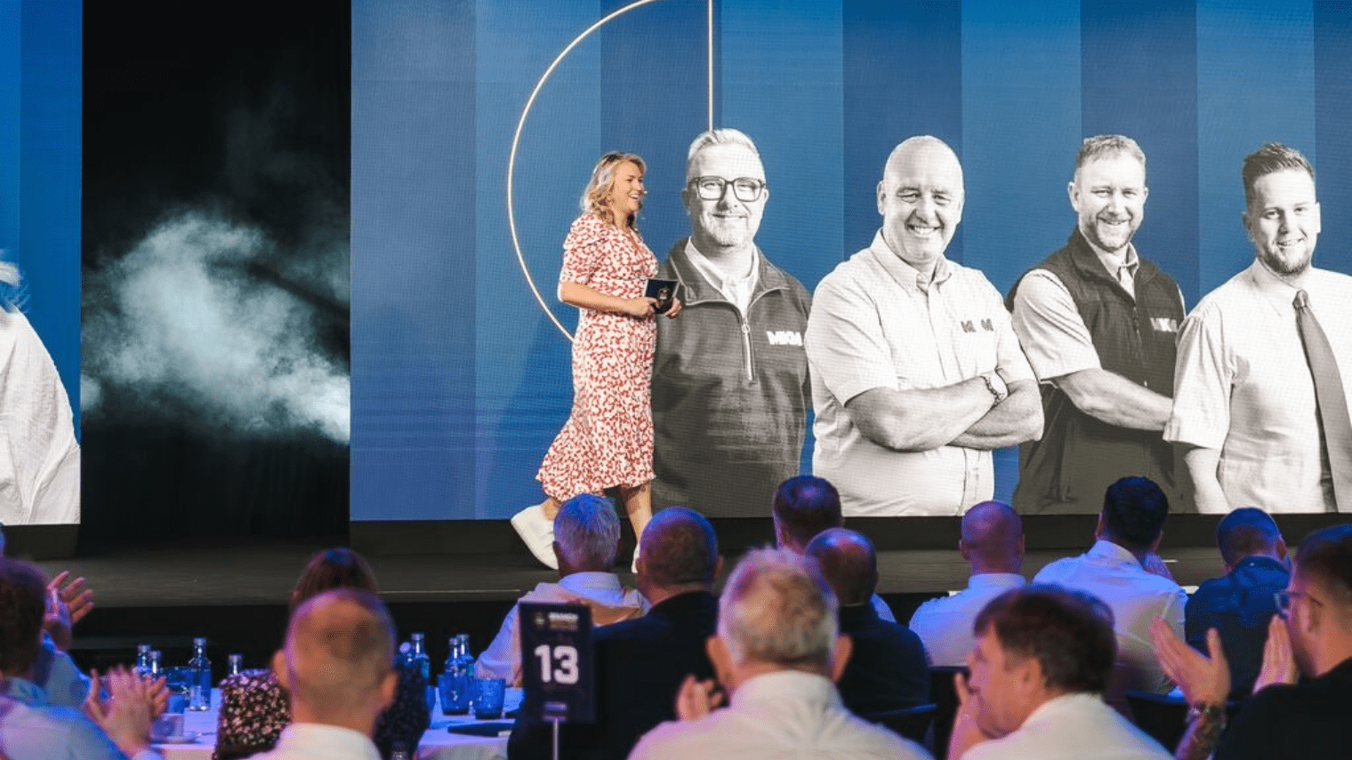At a strategic conference, a woman in a red and white dress walks onstage to applause. Behind her, a large screen displays black-and-white images of four smiling men standing side by side.
