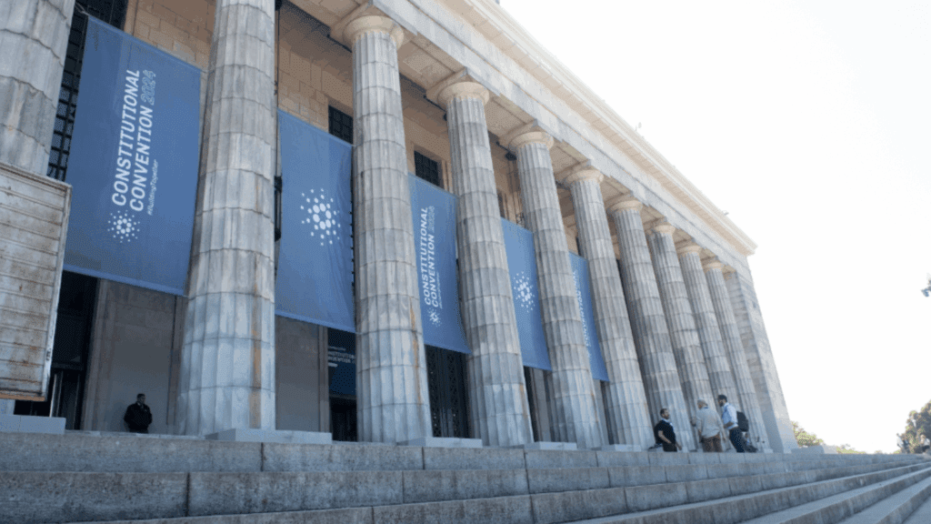 A neoclassical building with tall columns displays blue banners reading Constitutional Convention. Several people, possibly attendees of a blockchain event, are standing or sitting on the steps in front of the entrance.
