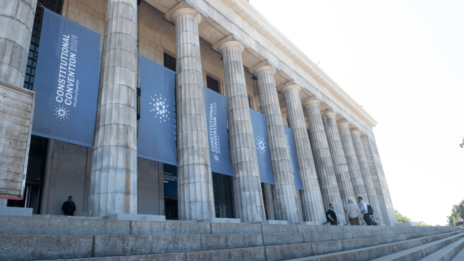 A neoclassical building with tall columns displays blue banners reading Constitutional Convention. Several people, possibly attendees of a blockchain event, are standing or sitting on the steps in front of the entrance.