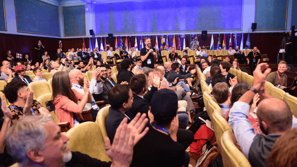 A large group of people sits in an auditorium with yellow seats, clapping and smiling as one person stands and speaks at a lively blockchain event. Numerous national flags line the stage at the front of the bright, energetic room.