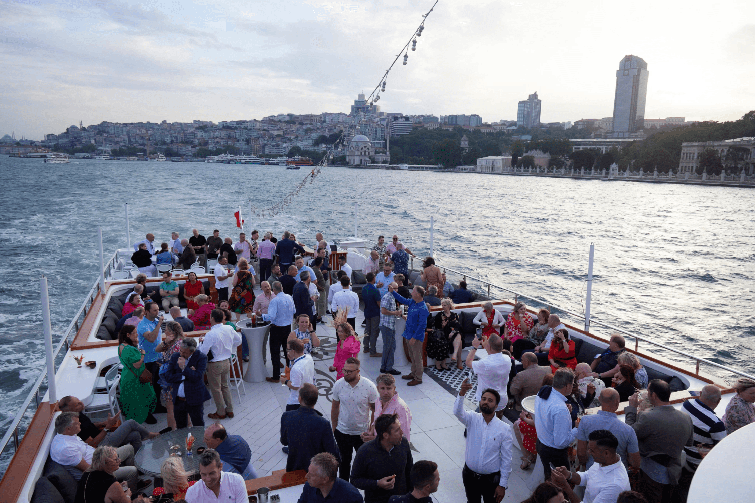 A large group of people socializes on the deck of a boat cruising along a city waterfront at sunset