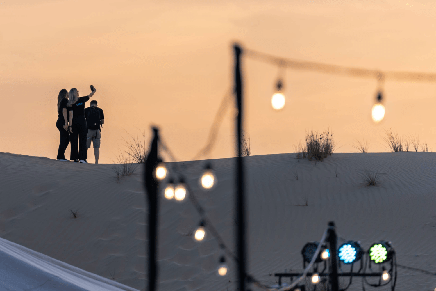 Three people stand on a sand dune at sunset, with one person taking a selfie. Out-of-focus string lights and stage lights are visible in the foreground, adding a warm, festive atmosphere.