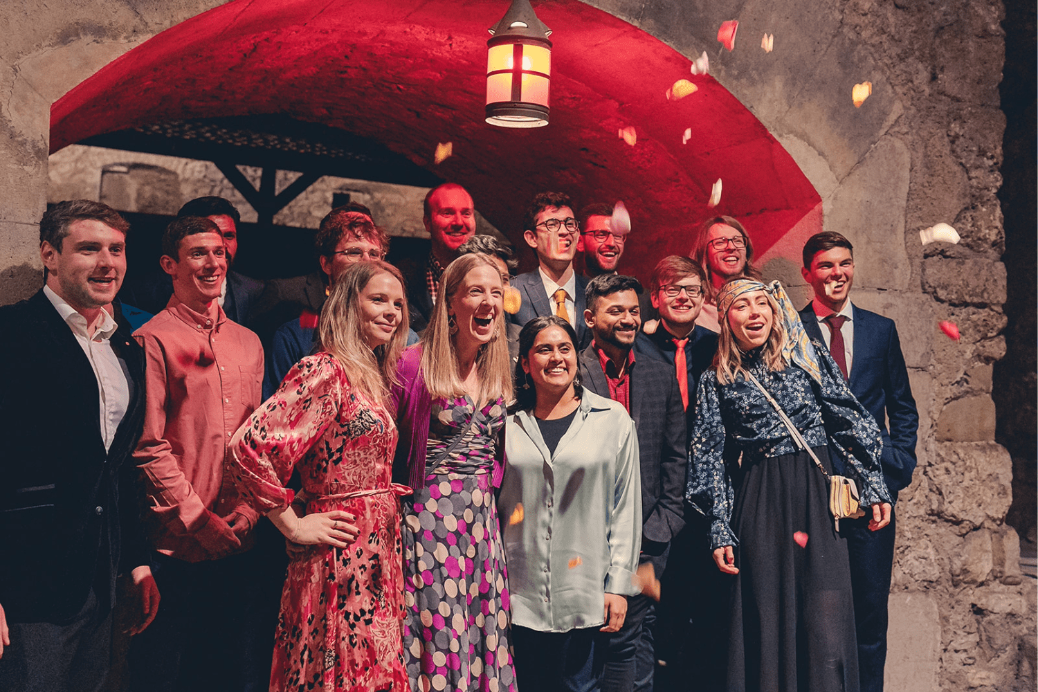 A group of people dressed in formal and semi-formal attire stand closely together, smiling and laughing under a red-lit stone archway as rose petals fall around them, beautifully tackling loneliness through joyful connection.