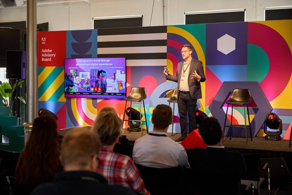 A man in business attire speaks on stage at an Adobe Advisory Board event, enhanced by creative event services, with a colorful geometric backdrop and a screen displaying a digital experiences presentation to an audience.