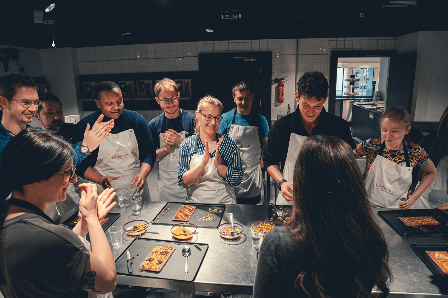 A group of people wearing aprons stand around a kitchen counter, smiling and clapping as they admire finished desserts on plates during a cooking class or workshop.
