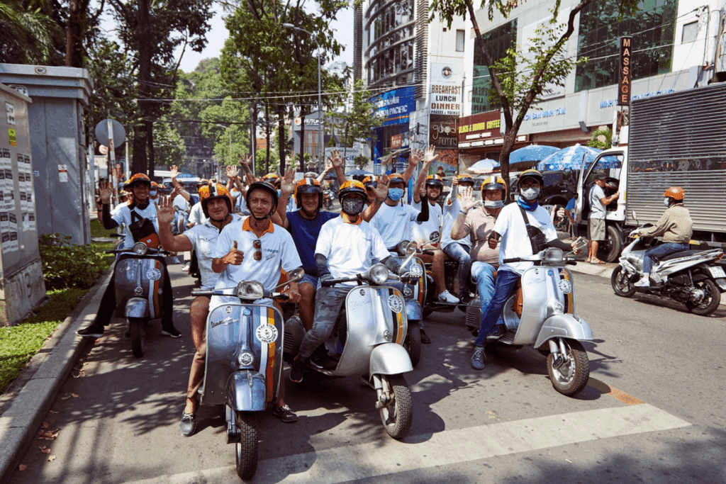 A group of people wearing helmets sit on scooters, smiling and giving thumbs up during an Incentive Events outing, on a city street lined with trees and buildings on a sunny day.
