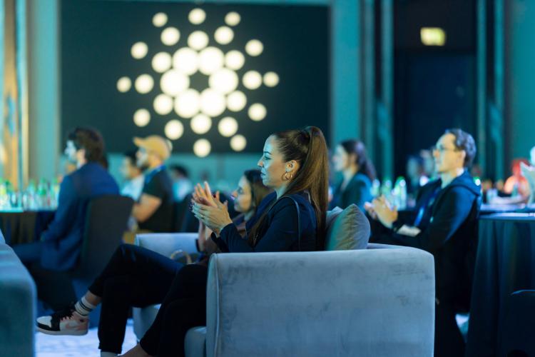 People sit on sofas and chairs, attentively watching and applauding at an indoor event enhanced by creative event services. The background features circular lights on a dark wall, with tables set with water bottles in this modern, professional atmosphere.
