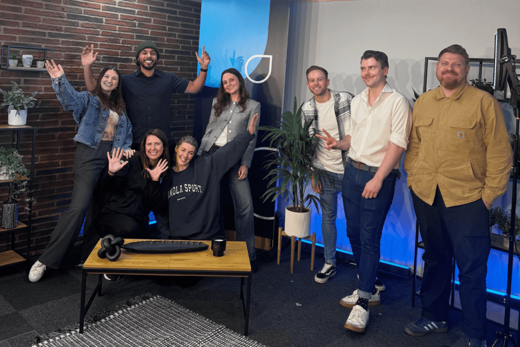 Eight people pose and smile in a modern studio space with brick and blue walls, plants, and recording equipment. Several wave enthusiastically, creating a joyful, casual group atmosphere—capturing the excitement of their About First Event.
