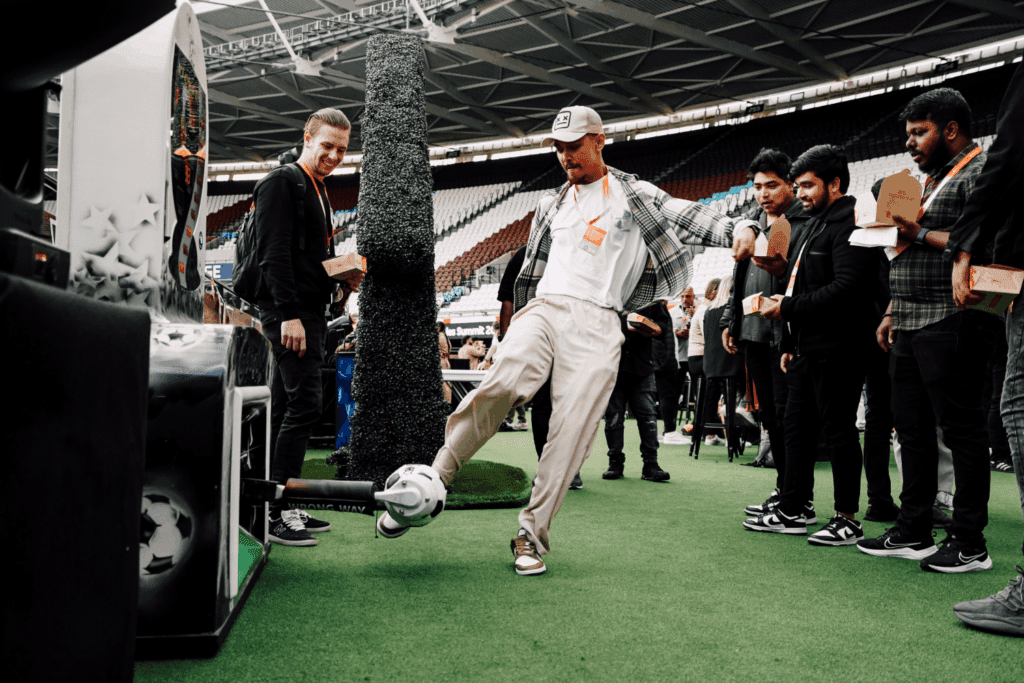 At an Experiential Event, a man kicks a soccer ball on artificial turf indoors while others watch, some holding food boxes. The lively scene unfolds in a stadium-like venue, capturing the excitement and energy of the moment.
