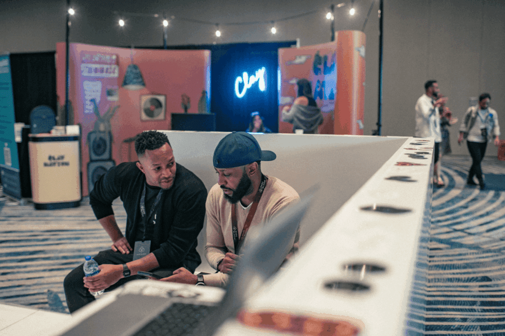 Two men sit and talk at an indoor experiential event with colorful booths and a neon “Clay” sign in the background. Other people are visible standing and walking around the lively venue.