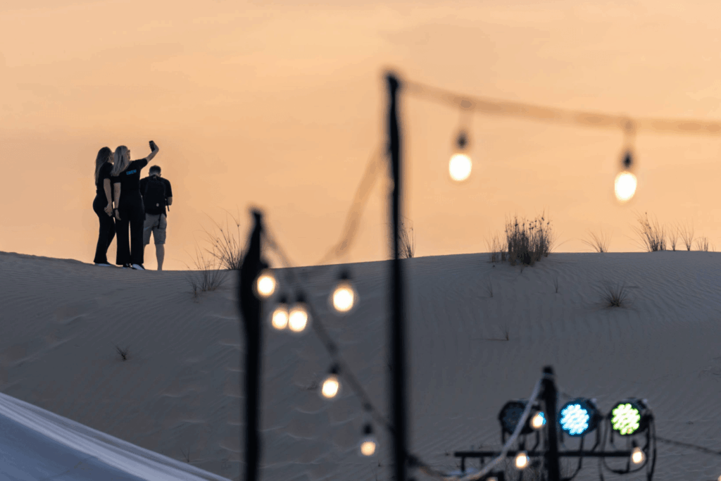 Three people stand on a sandy dune at sunset, one holding up a phone for a selfie. Out-of-focus string lights and stage lights in the foreground hint at vibrant Event Marketing & Communications happening under the soft orange sky.