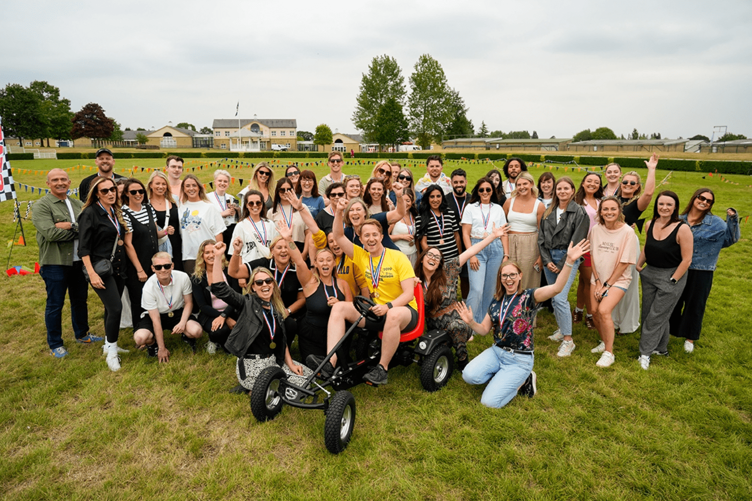 A large group of people pose cheerfully outdoors on grass at an event careers gathering, surrounding a person in a yellow shirt sitting on a pedal go-kart. Trees, buildings, and a racetrack appear in the background under a cloudy sky.