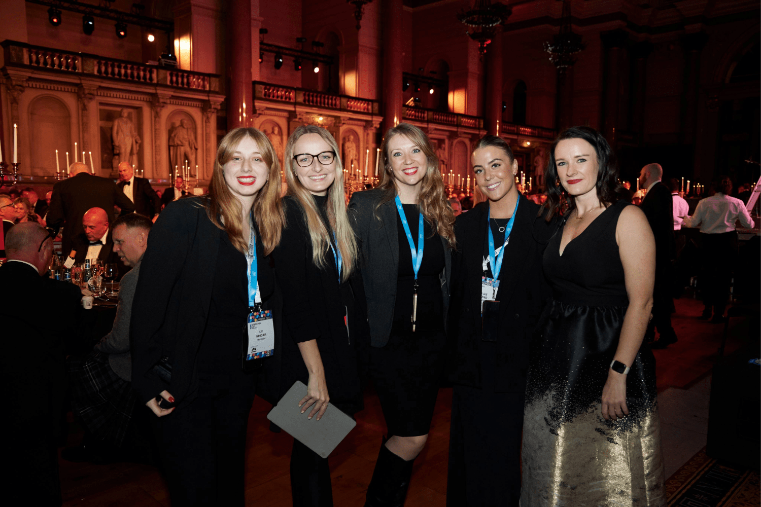 Five women dressed in formal attire smile for a group photo at a gala event, celebrating their achievements in event careers amid candlelit tables and ornate architecture in the grand hall.