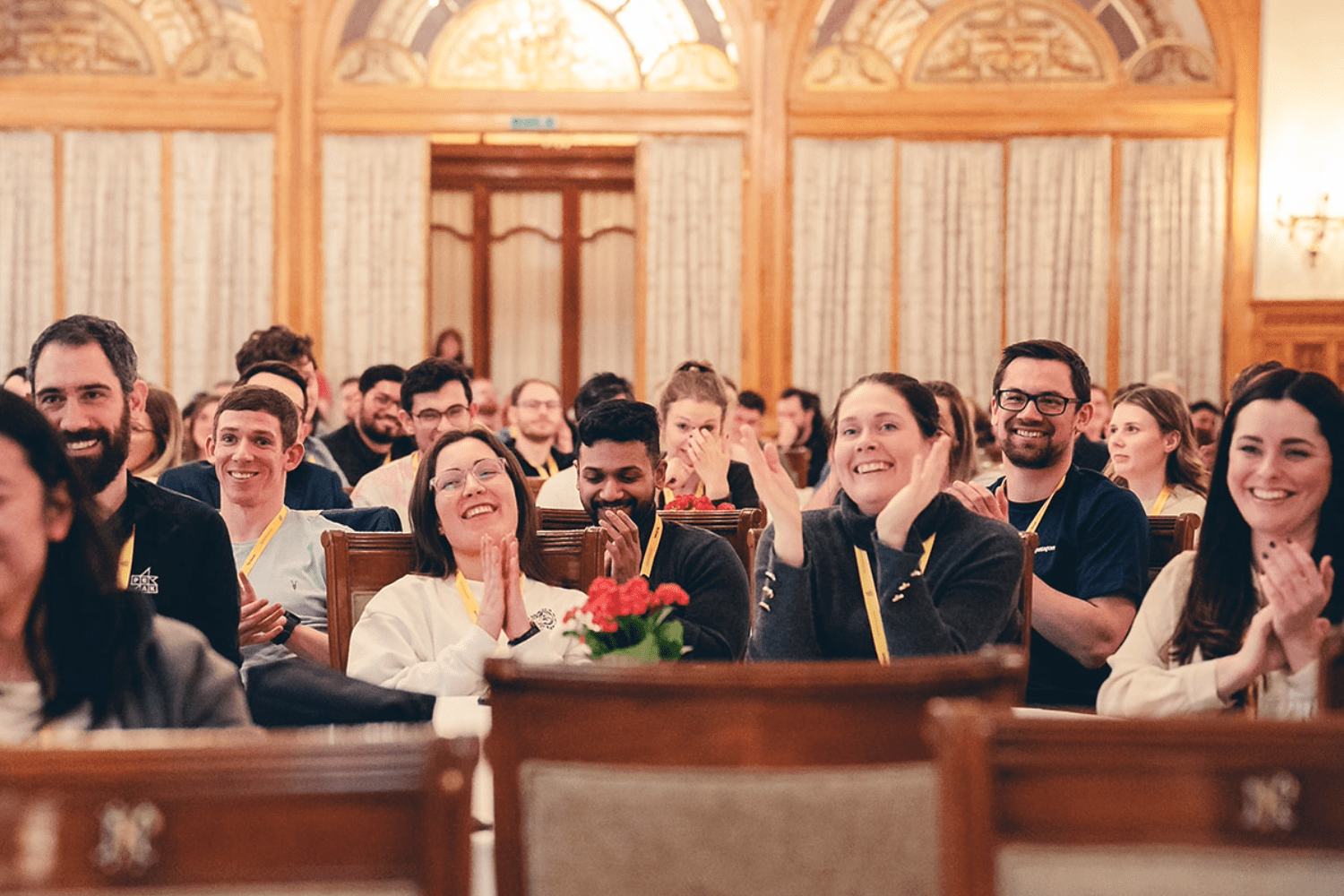 A diverse group, including members of hybrid teams, sit in rows inside an ornate room, smiling and applauding. Some wear name tags and ribbons, and flowers decorate a table in the foreground. The atmosphere appears joyful and celebratory.