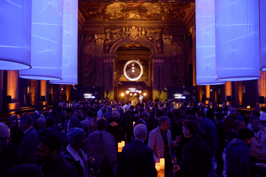 A large crowd gathers in an ornate, dimly lit hall with high ceilings and blue lighting. Decorative columns and a grand mural set the stage for unforgettable Event Experiences in this elegant, festive atmosphere.