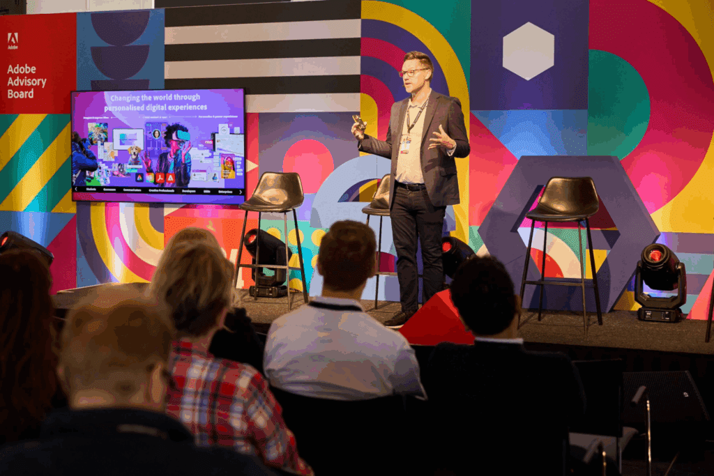 A man in business attire speaks on stage at a first event services gathering, in front of a colorful geometric backdrop and screen displaying Changing the world through personalized digital experiences, to an audience seated in chairs.