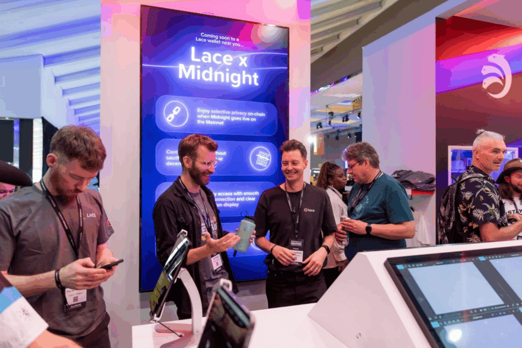 A group of people interact and smile at a tech expo booth with a large blue display reading Lace x Midnight, enjoying engaging Event Experiences. Digital devices and promotional materials are visible on the counter in front of them.