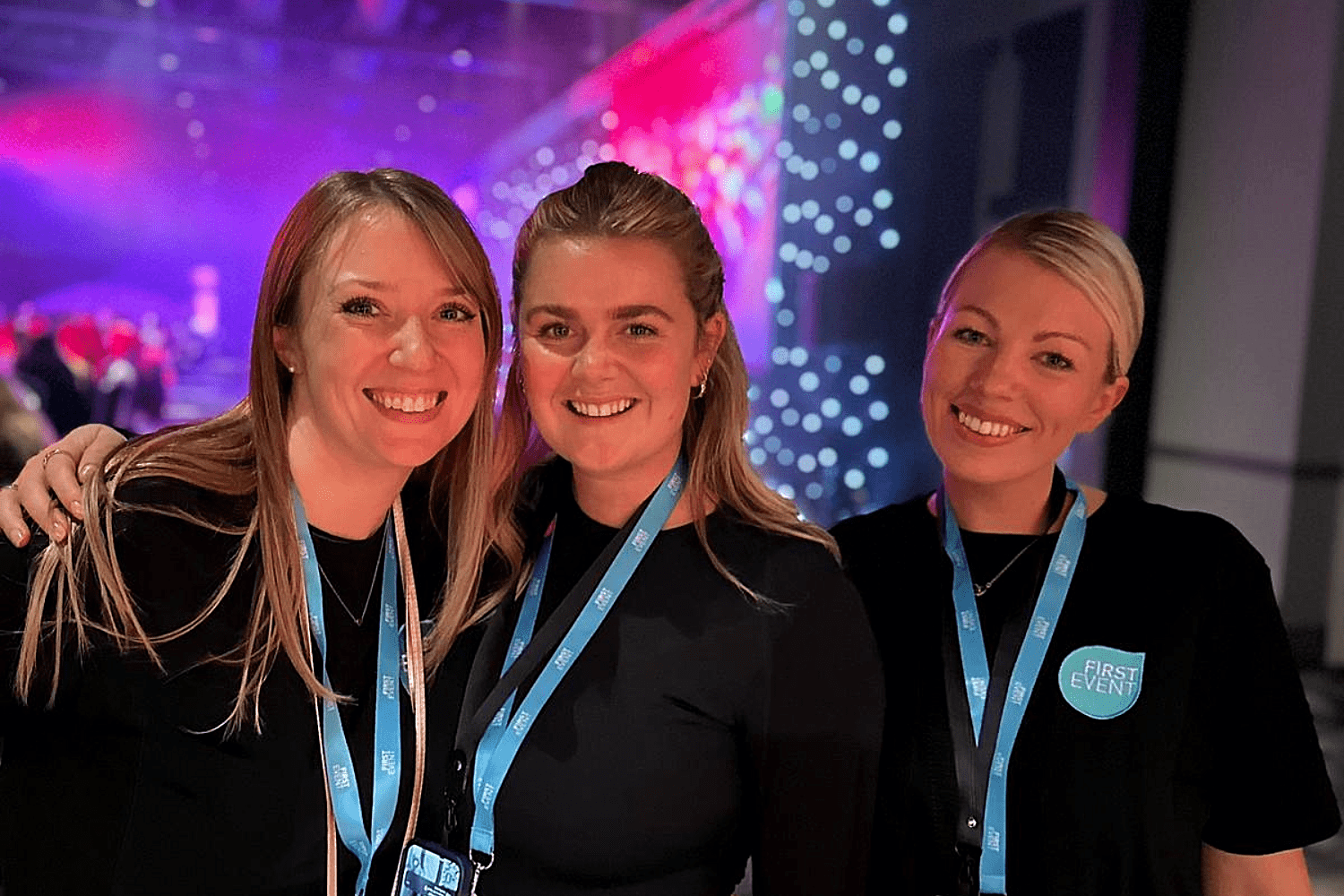 Three women wearing black shirts and blue lanyards with “FIRST EVENT” logos smile together at an indoor event, celebrating their passion for event careers amid colorful lights and a blurred background.