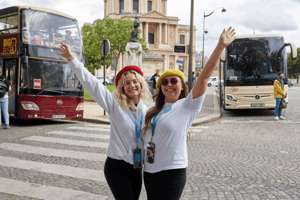 Two smiling women wearing berets and tour guide lanyards stand on a cobblestone street with arms raised, showcasing their passion for event careers in front of tour buses and a historic building in a lively city setting.
