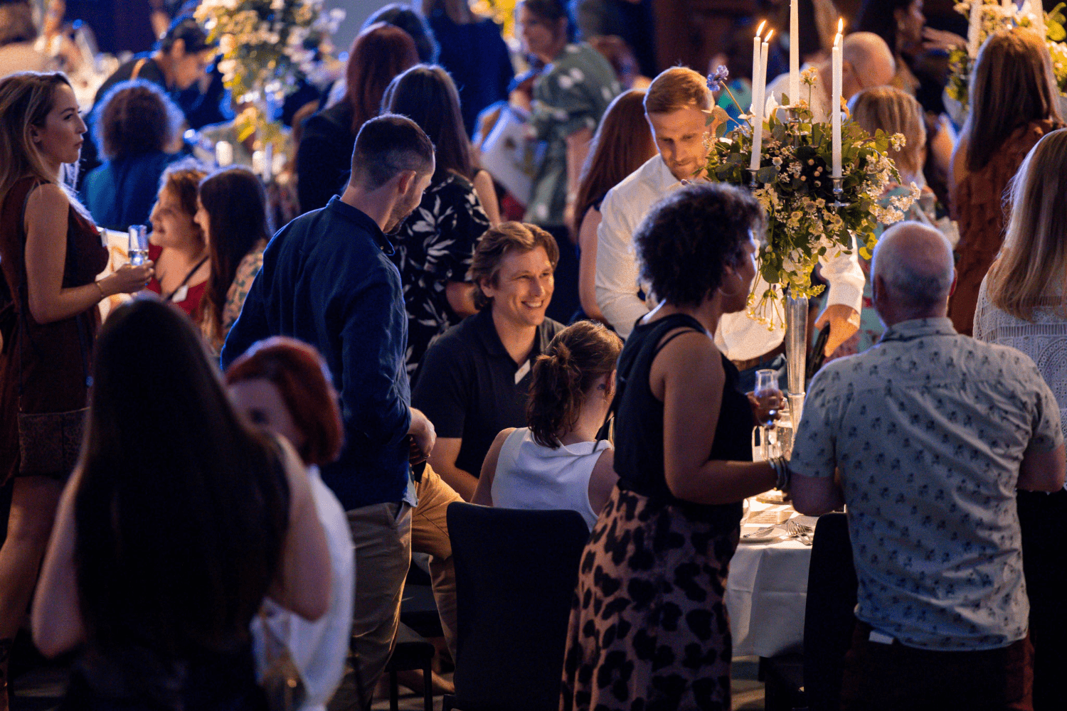 A lively indoor event focused on employee engagement, with people socializing around tables decorated with flowers and candles. Some guests are standing, others are seated, and the atmosphere is warm and festive.