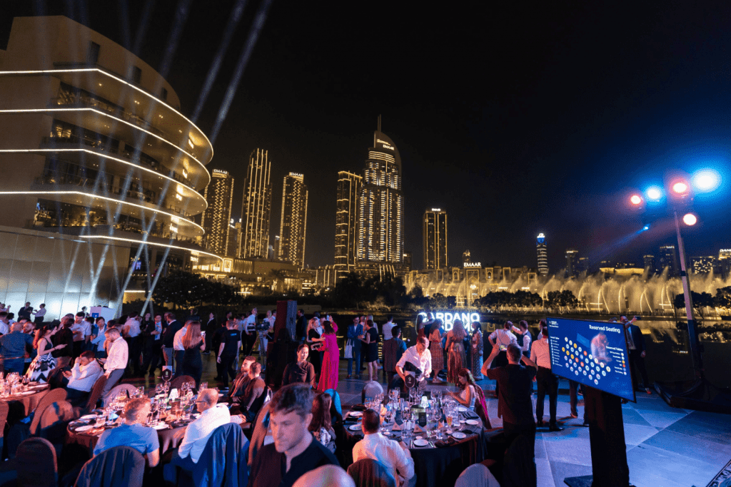 An outdoor evening event with people dining at round tables, city skyscrapers and lights in the background, a fountain, and a large screen displaying information—perfect for an event space finder. Spotlights illuminate the scene.