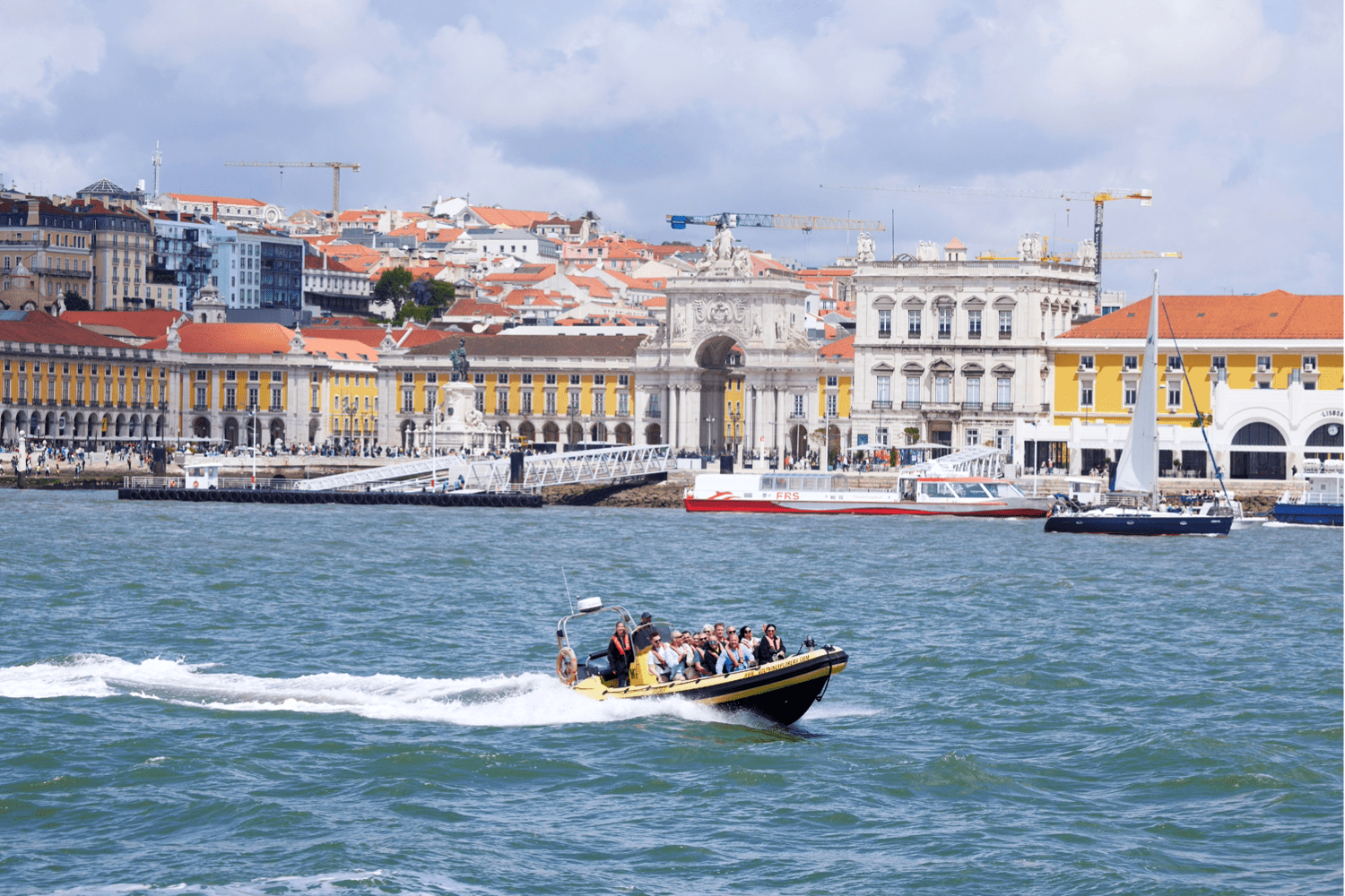 A small motorboat with people on board speeds across a river, with historic buildings, sailboats, and cranes visible along the waterfront in Lisbon, Portugal, under a partly cloudy sky.