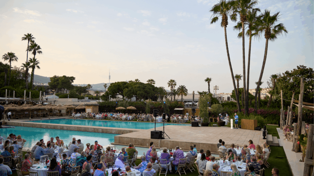 Outdoor industry conference by a pool, with many people seated at round tables; palm trees sway in the background and a stage with speakers is set up near the water under a partly cloudy sky.