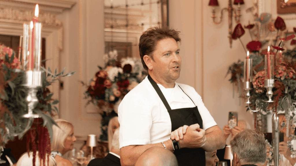 A chef in a white shirt and black apron speaks to seated guests at an elegant, ornately decorated dining event with candles and floral arrangements, creating a refined atmosphere reminiscent of a strategic conference dinner.