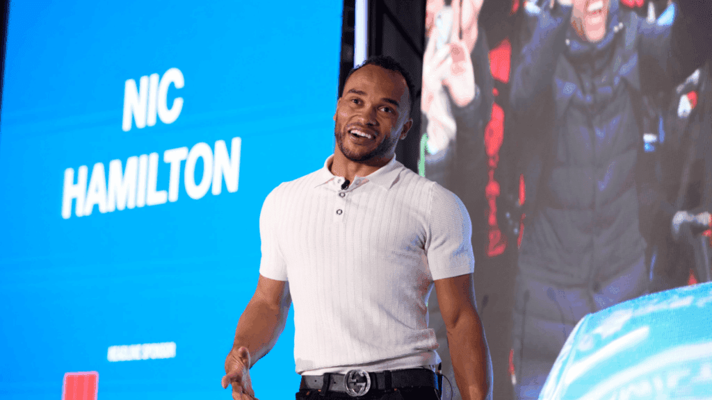 A man wearing a white short-sleeve shirt stands and smiles on stage at an industry conference, in front of a large blue screen displaying the name Nic Hamilton.