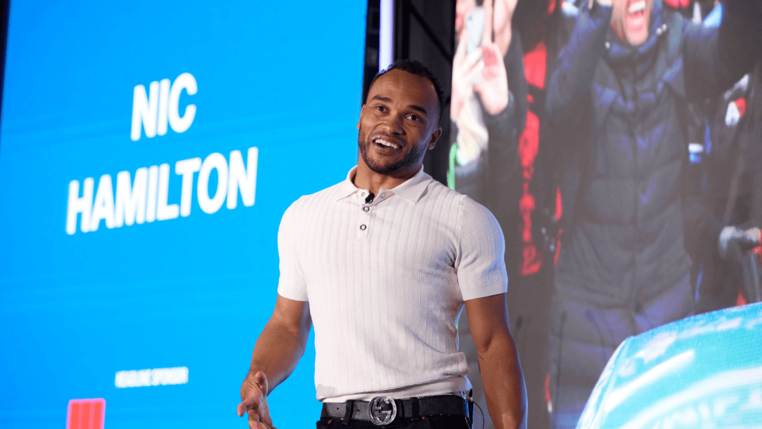 A man wearing a white short-sleeve shirt stands and smiles on stage at an industry conference, in front of a large blue screen displaying the name Nic Hamilton.