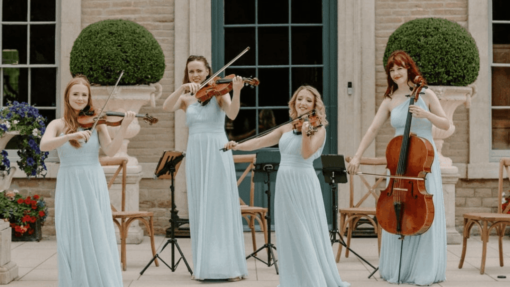 Four women in matching light blue dresses perform outdoors at a strategic conference, three playing violins and one on cello, standing before a building with large windows and decorative topiary bushes.