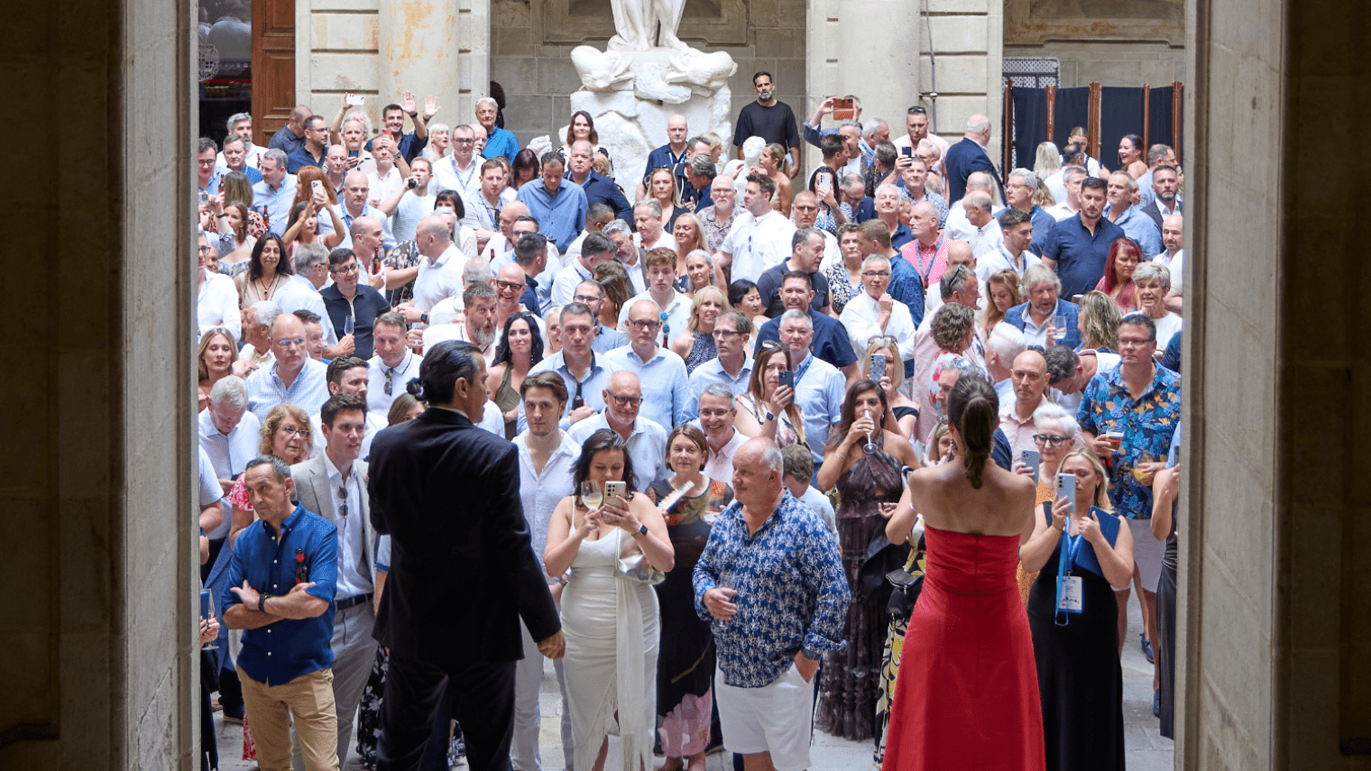 A large crowd of people, many dressed in white or light clothing, gathers in a courtyard with stone walls and a statue for an industry conference. Several attendees are taking photos or videos while two people stand at the front facing the crowd.