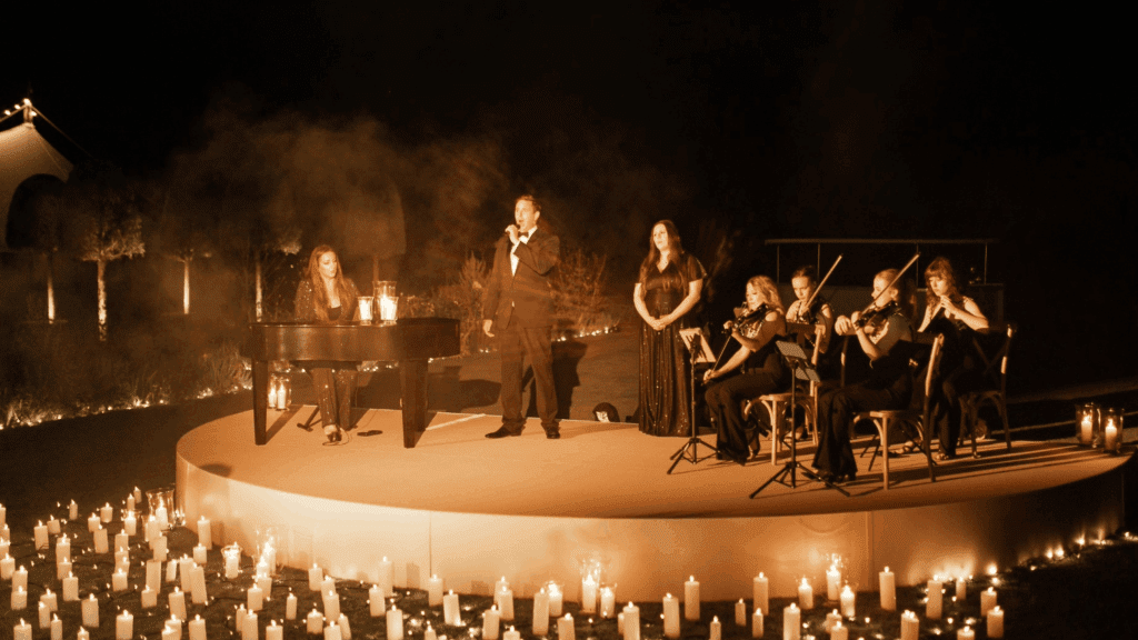 A group of musicians in formal attire perform on a round, candlelit outdoor stage at night, creating an ambiance as elegant as a strategic conference, with a pianist, singer, and string ensemble surrounded by glowing white candles.