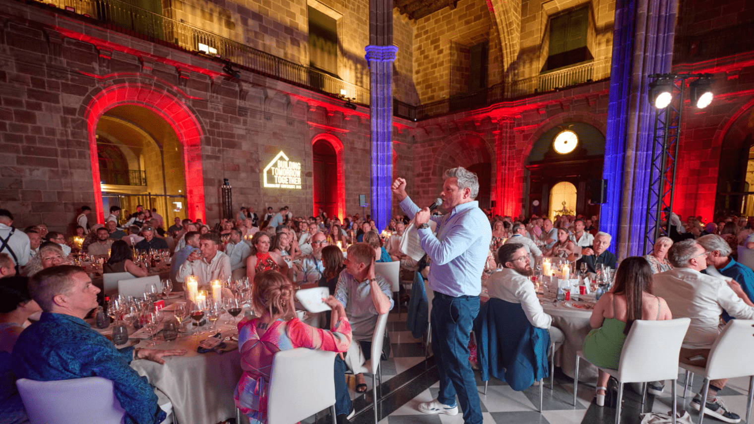 A man stands and speaks with a microphone to a large crowd at an industry conference, seated at round tables in a grand, historic hall with high arched ceilings, dramatic lighting, and stone walls.