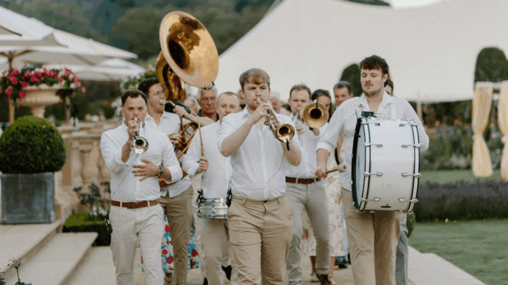 A brass band dressed in white shirts and beige pants marches outdoors at a strategic conference, playing drums, trumpets, and a sousaphone, with tents and greenery visible in the background.