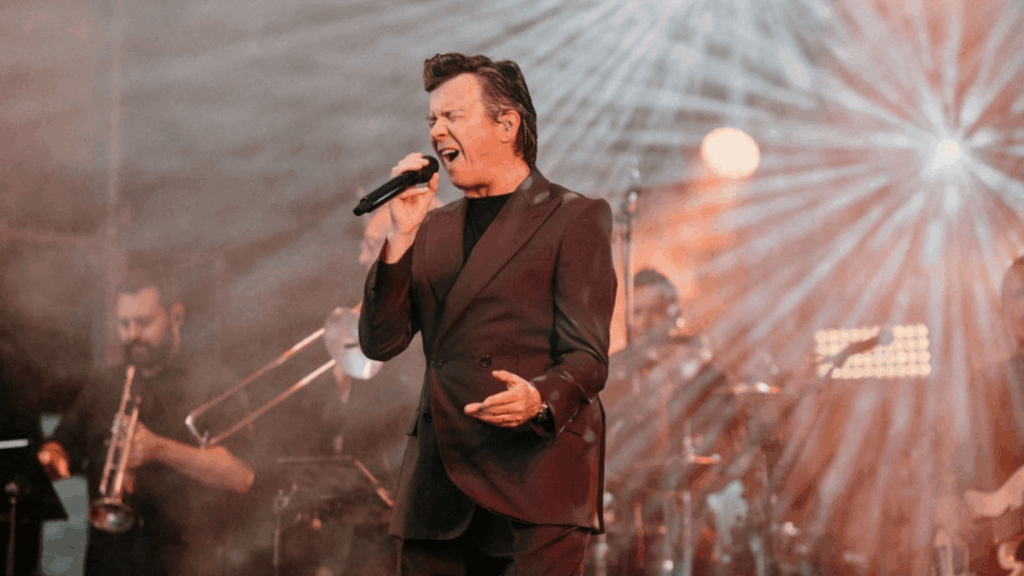 A male singer in a dark suit passionately performs on stage with a microphone, surrounded by band members and dramatic stage lighting, as if energizing the crowd at a strategic conference.