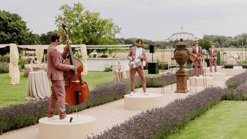 A band of four men in matching pink suits performs outdoors at a strategic conference, each standing on a circular platform amid lavender bushes, playing upright bass, drums, guitar, and saxophone in a garden setting.