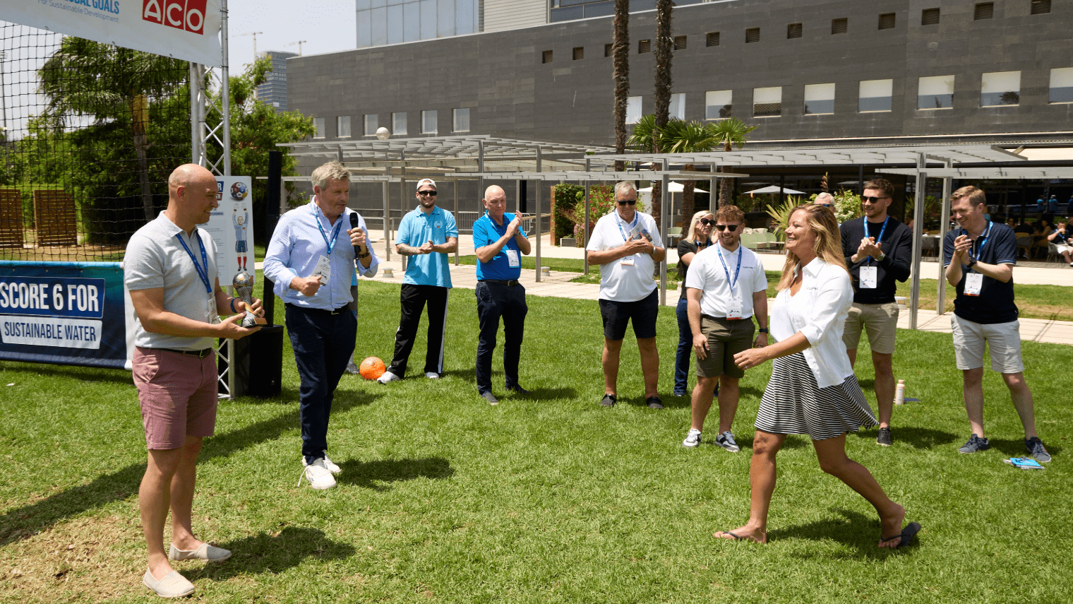 A group gathers outdoors on a grassy field for their About First Event. A man with a microphone stands near a woman walking toward him, while others watch, clap, and smile. There is a soccer goal and a banner in the background.