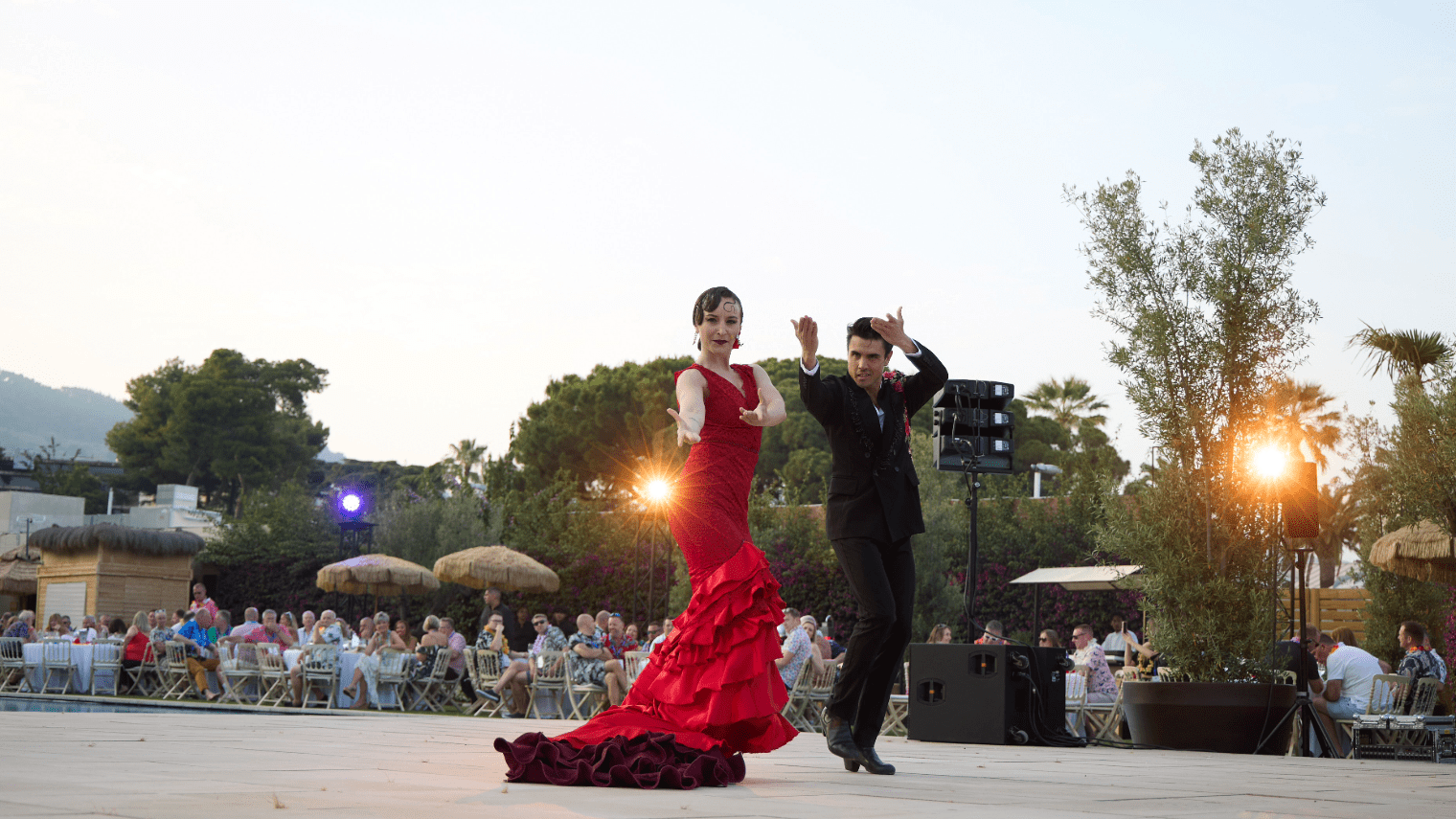 A woman in a red flamenco dress and a man in a black suit perform an outdoor dance at sunset during the About First Event, with an audience seated at tables and lush greenery in the background.