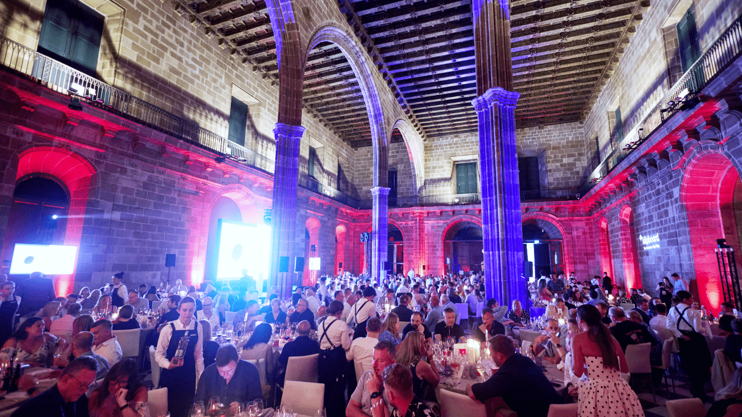A large formal event, reminiscent of About First Event, unfolds in a grand hall with stone arches and columns. Seated guests at round tables bask in blue and red lights that illuminate the historic venue, creating an elegant atmosphere.