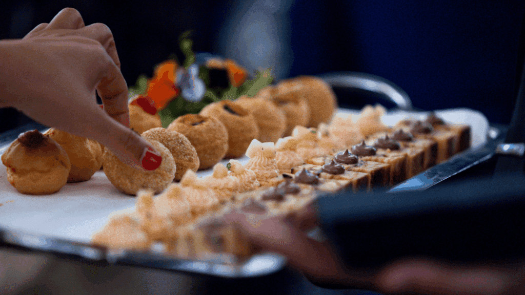 At a private luxury event, a hand with red nail polish reaches for a pastry from a tray filled with assorted desserts, including cream puffs and small cakes, as another person holds the tray in a dimly lit setting.