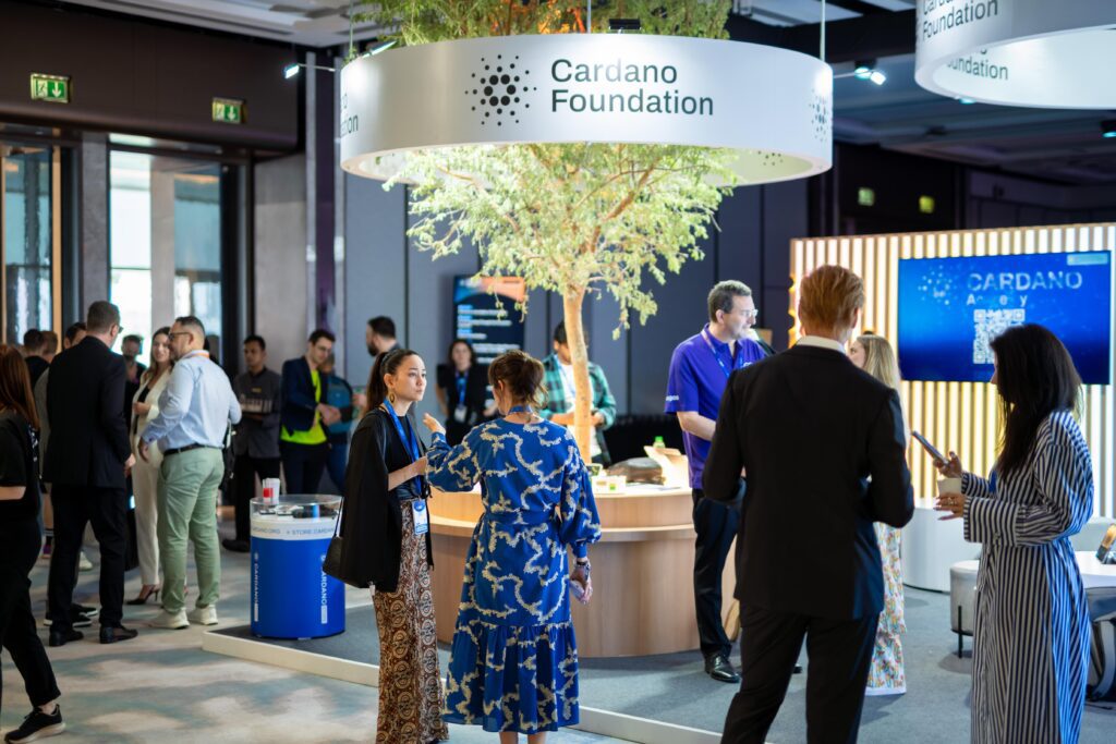 People gather and converse around the Cardano Foundation booth at a conference, featuring a tree centerpiece and informational displays—an example of strategic event planning. Some attendees are engaged in discussion, while others explore the exhibit.