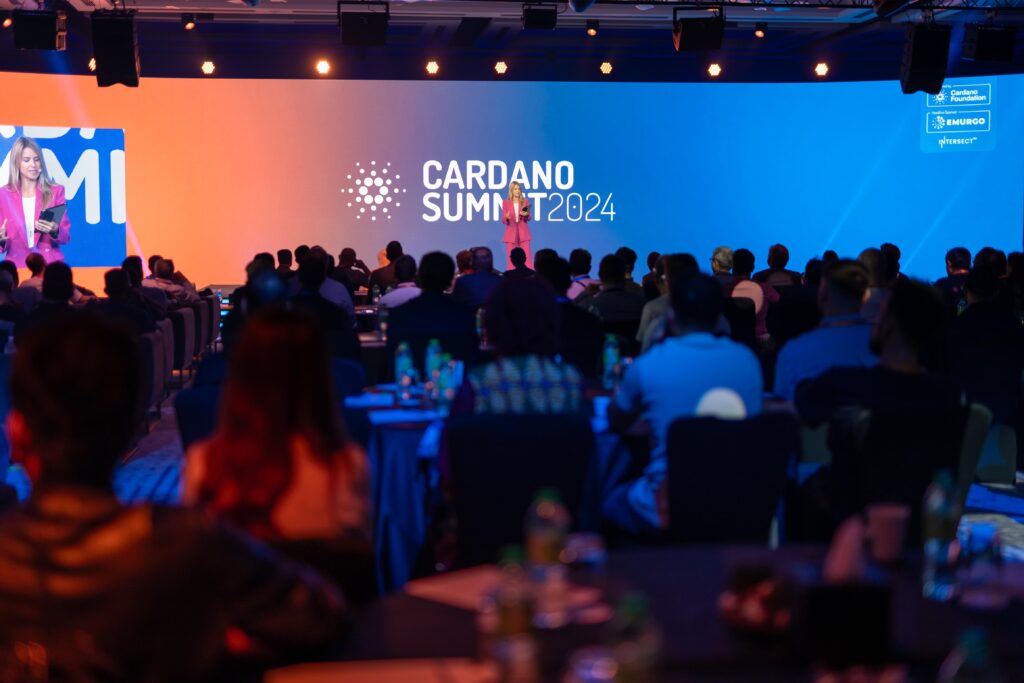 A large audience sits facing a stage with a bright screen displaying “Cardano Summit 2024.” As part of strategic event planning, tables are set up with water bottles and papers in the dimly lit room while a speaker addresses the crowd.