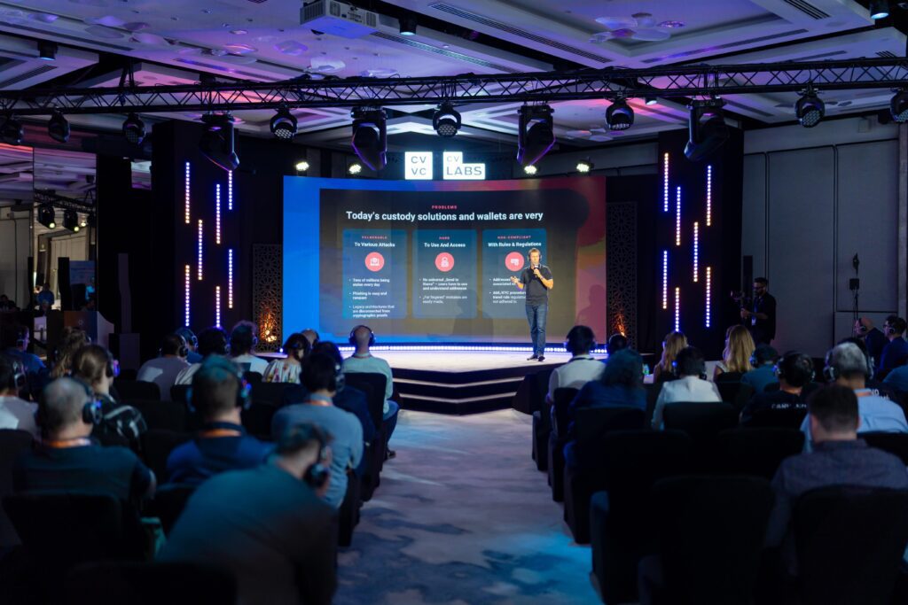 A speaker stands on stage presenting creative event services to an attentive audience in a conference room, with a large screen behind displaying information about custody solutions and wallets.