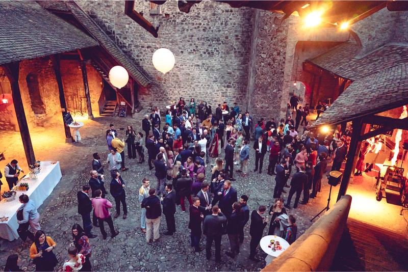 A large group of people socialize at an evening event in a rustic stone courtyard, enjoying unique event experiences with tables of food and drinks, warm lighting, and glowing white balloons creating a festive atmosphere.
