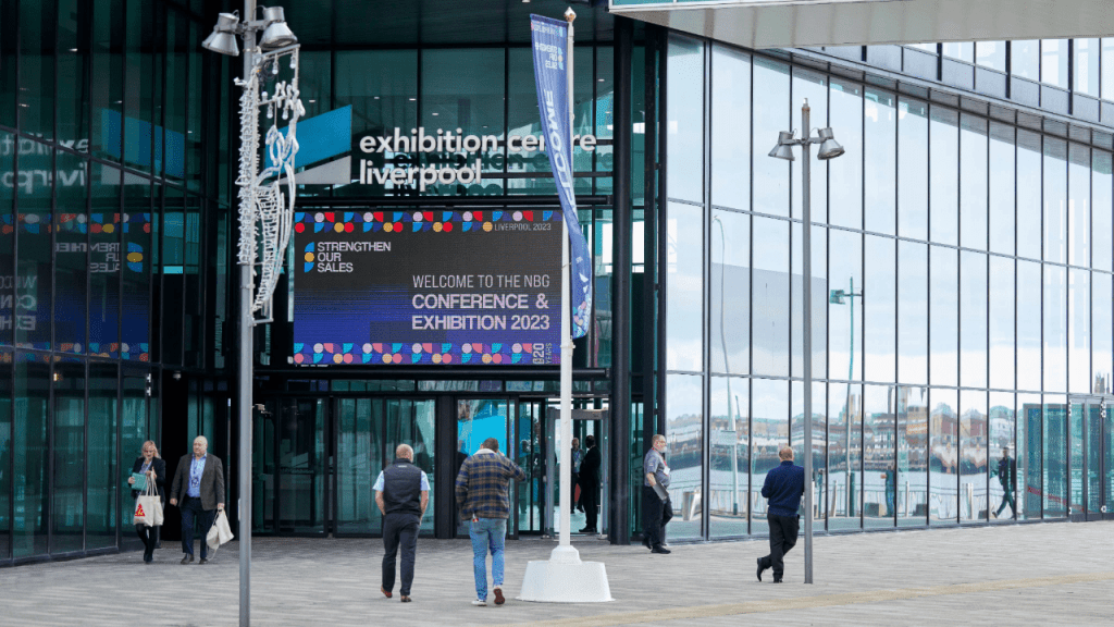 People walk outside the glass-walled Exhibition Centre Liverpool, a top choice for event space finder users. A large digital sign reads Welcome to the NBG Conference & Exhibition 2023. The weather appears clear and bright.