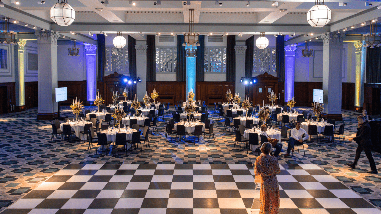 A large, elegant banquet hall with round tables set for an event. The floor has a black-and-white checkered pattern, and guests are mingling near the tables. This stunning venue, ideal for any occasion, was easily found using an event space finder.
