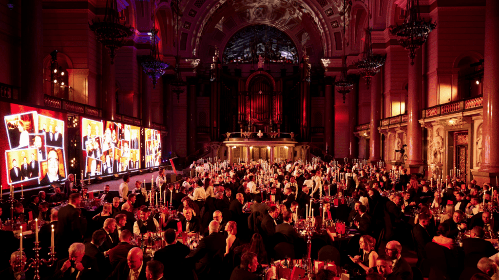 A grand, formal banquet in an ornate hall with red lighting. Attendees in evening wear sit at candlelit tables. Large screens display images, and a stage is set beneath a high, decorated ceiling—perfect for those seeking an event space finder.